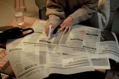 An adult woman marks job listings in a newspaper while sitting indoors, highlighting job search activity.