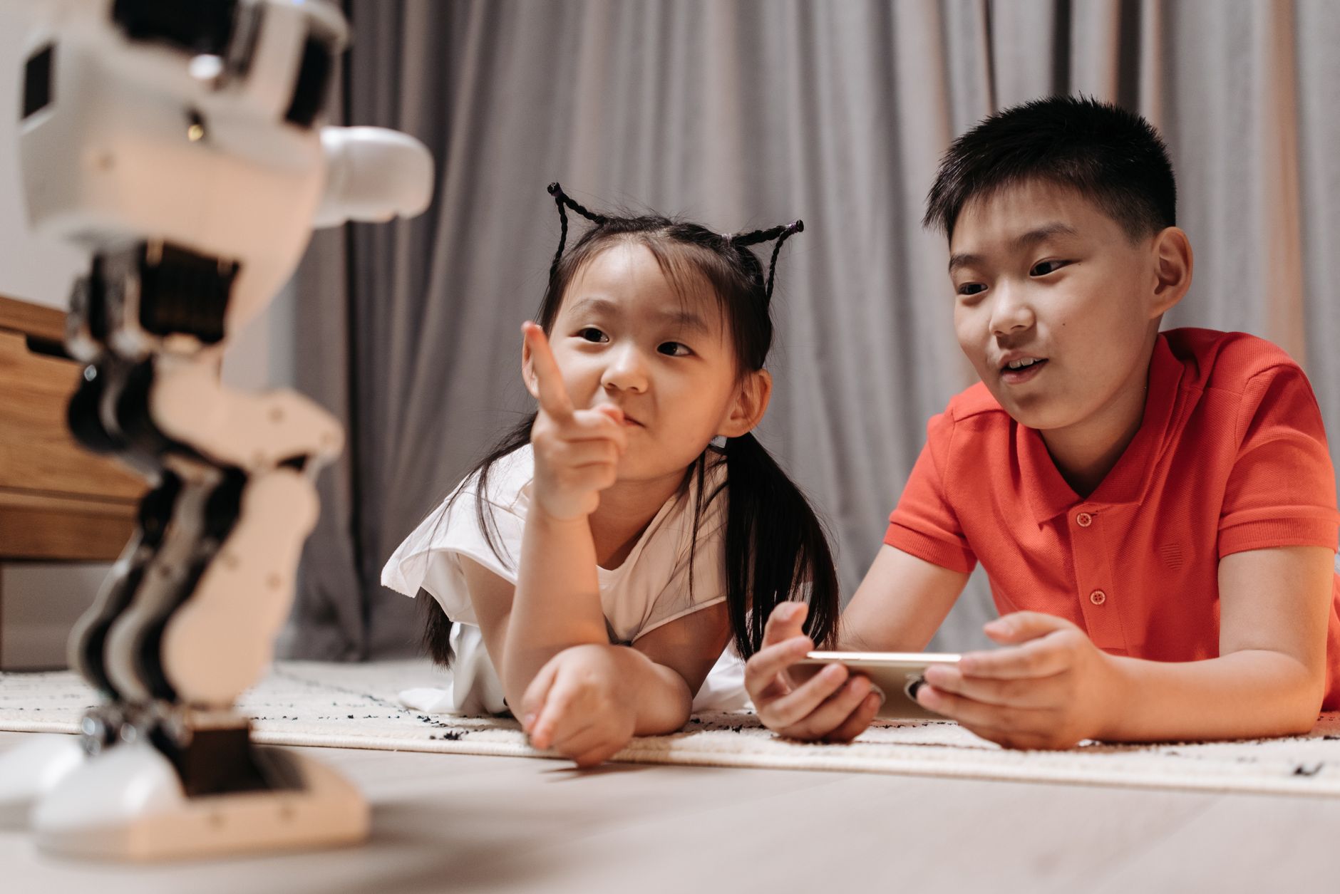 Two children interact with a robotic toy while using a smartphone, showcasing modern technology indoors.