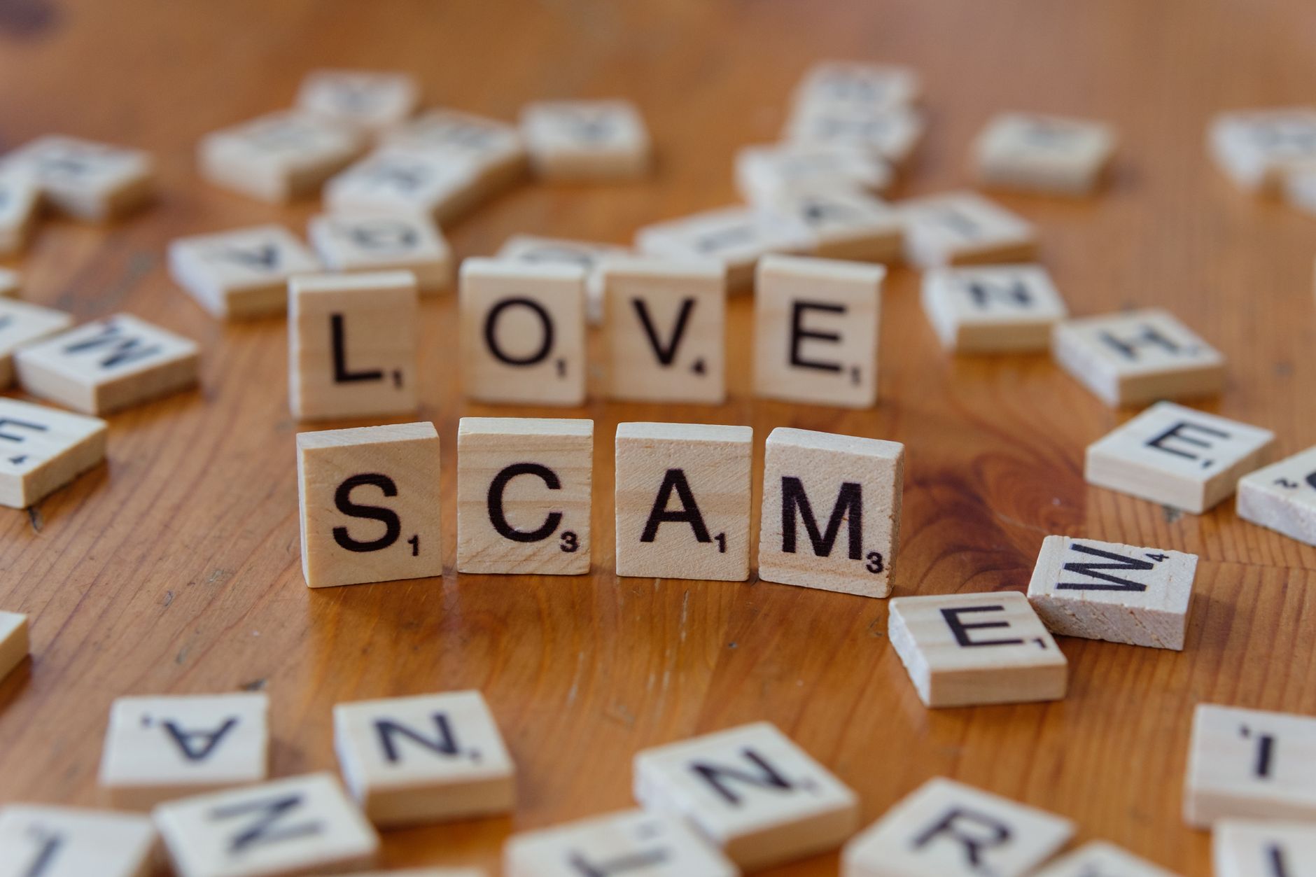 Scrabble tiles form words 'LOVE' and 'SCAM' on a wooden surface, illustrating deceit in relationships.