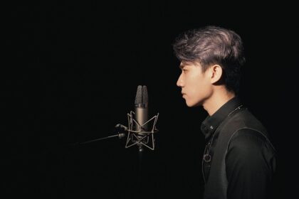 A young Asian male singer recording in a studio with a condenser microphone against a black background.