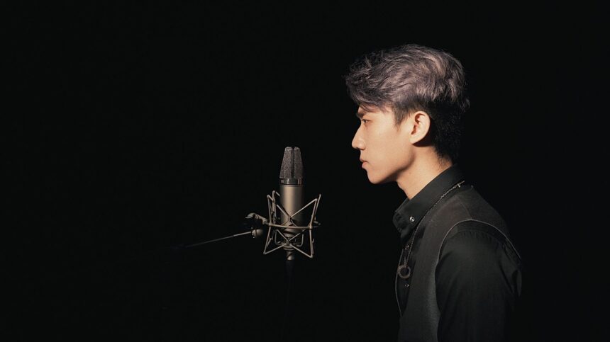 A young Asian male singer recording in a studio with a condenser microphone against a black background.