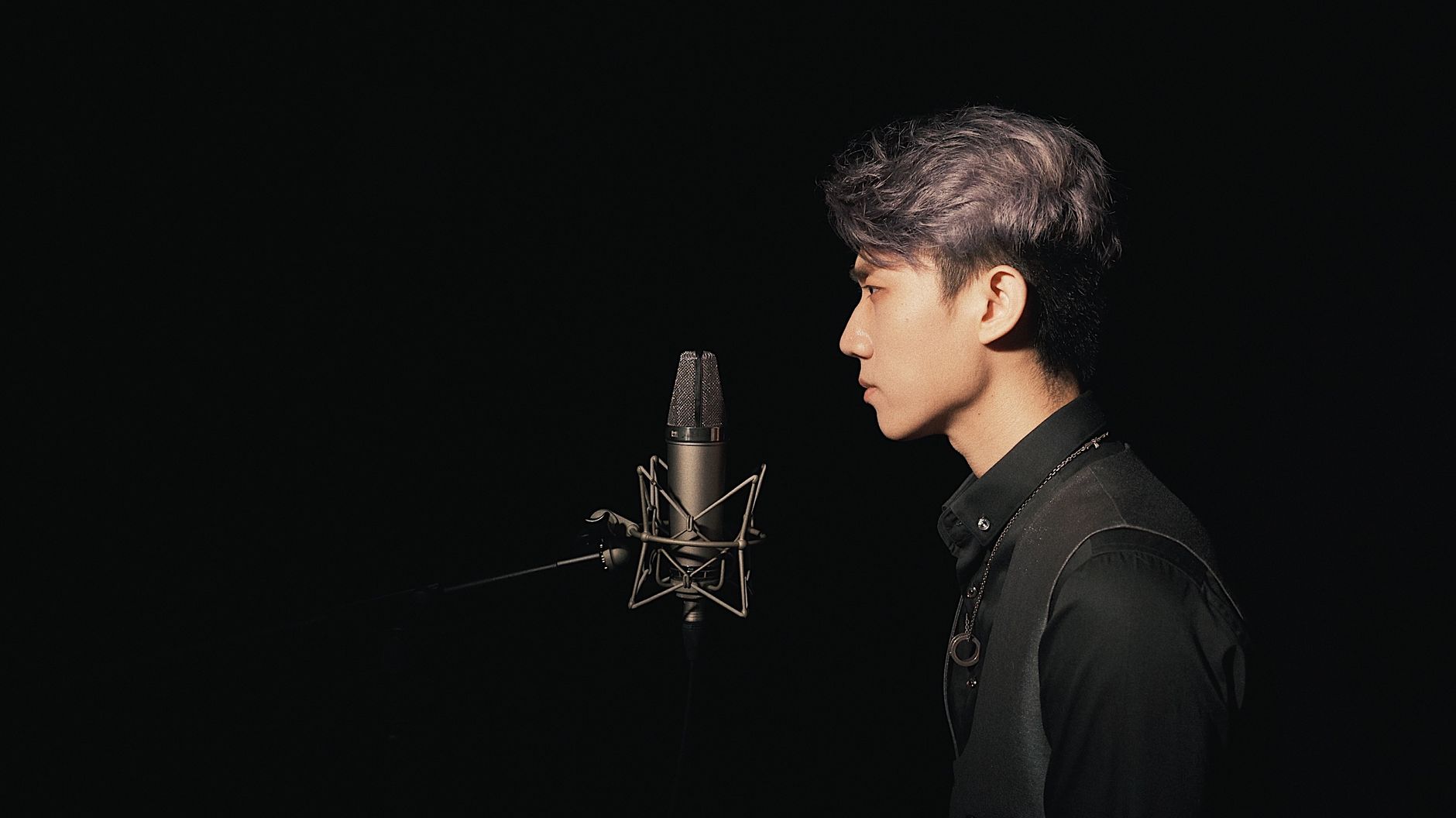 A young Asian male singer recording in a studio with a condenser microphone against a black background.