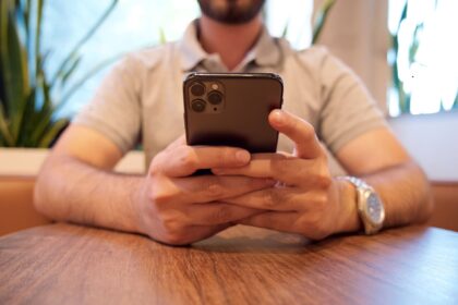 Close-up of a man holding a smartphone at a cafe table. Casual setting with plants in the background.