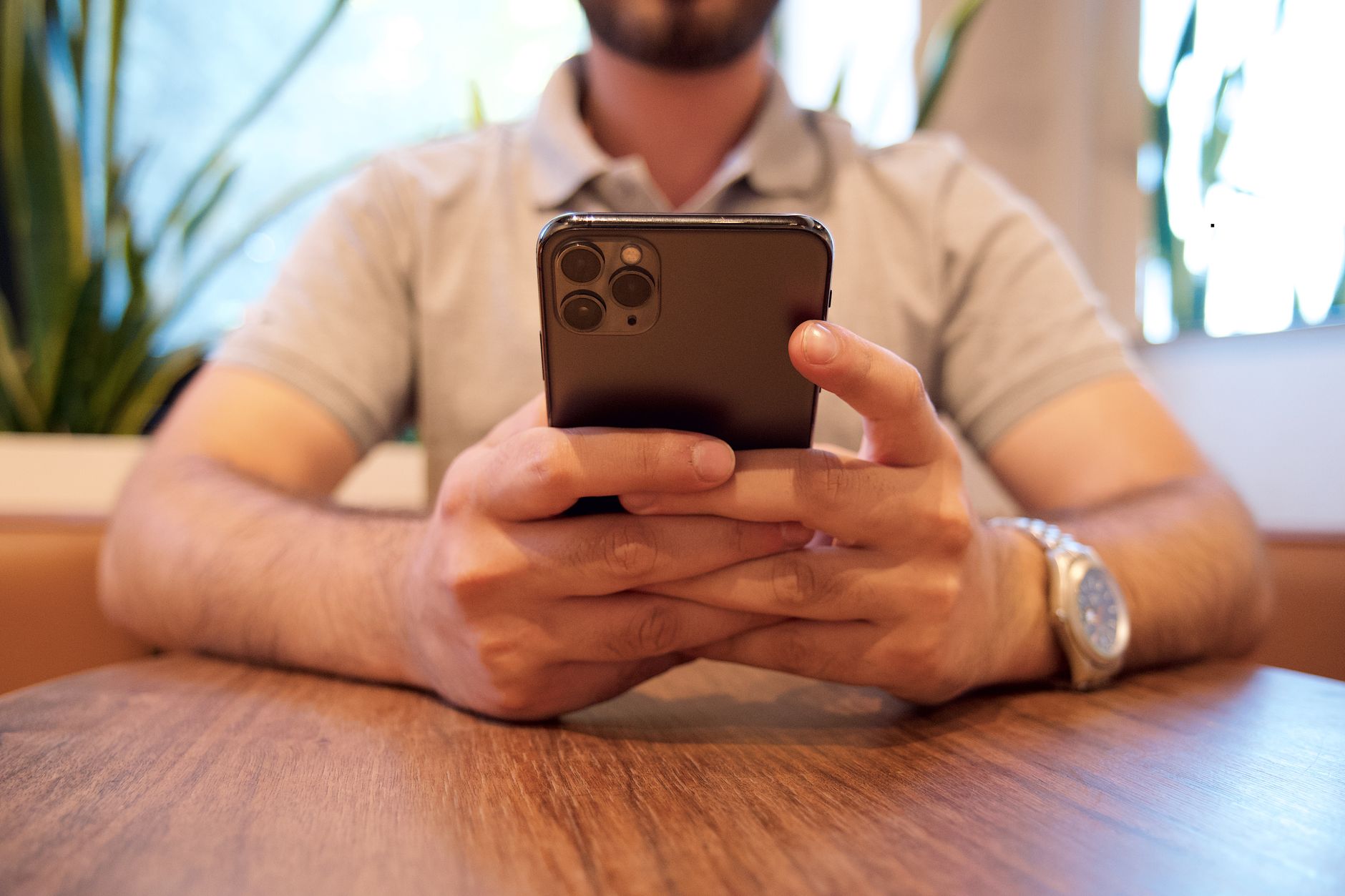 Close-up of a man holding a smartphone at a cafe table. Casual setting with plants in the background.