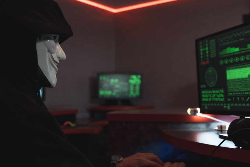 A person wearing a hacker mask operates a computer in a dimly lit room with digital displays.