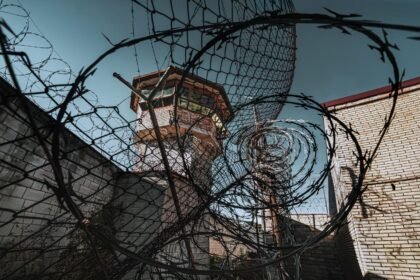 A dramatic view of a prison watchtower seen through coiled barbed wire under a clear sky.