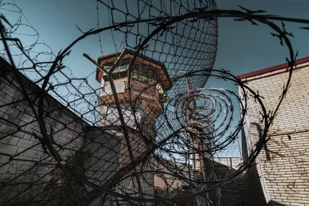 A dramatic view of a prison watchtower seen through coiled barbed wire under a clear sky.