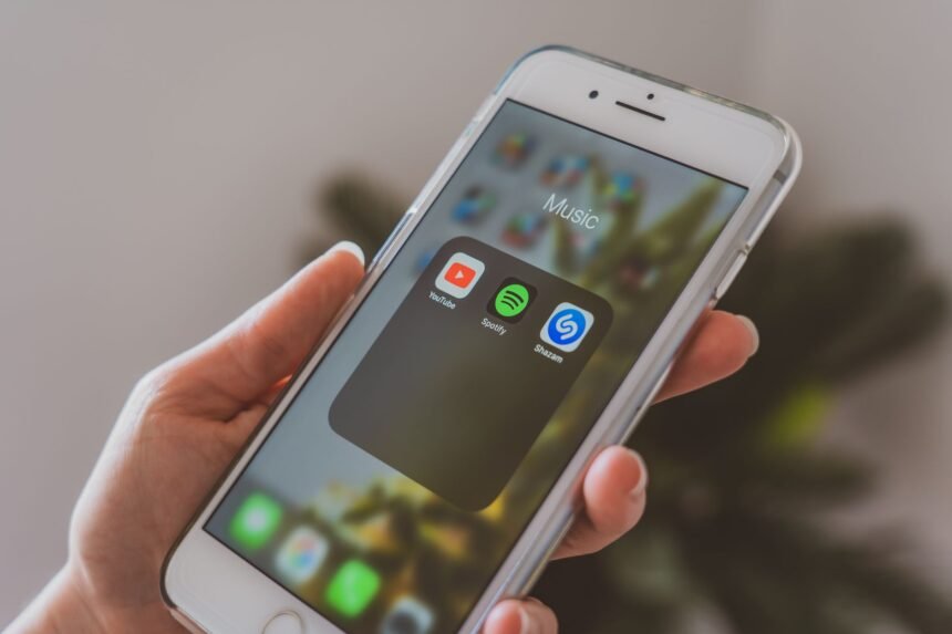 Hand holding a smartphone displaying popular music apps like Spotify and Shazam in a close-up shot.