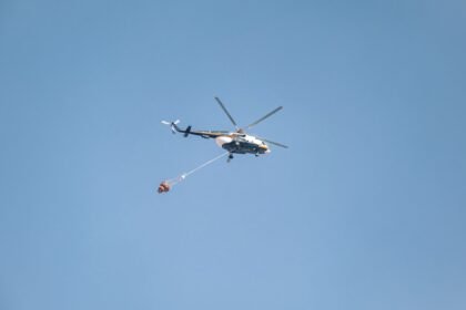 Helicopter with water bucket fighting a wildfire under a clear blue sky.
