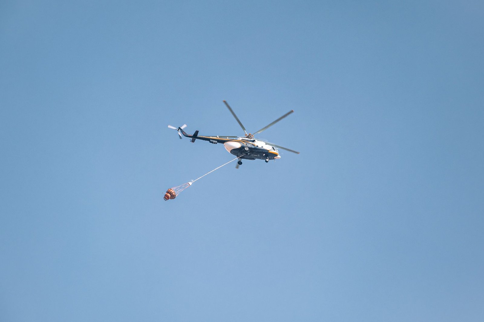 Helicopter with water bucket fighting a wildfire under a clear blue sky.