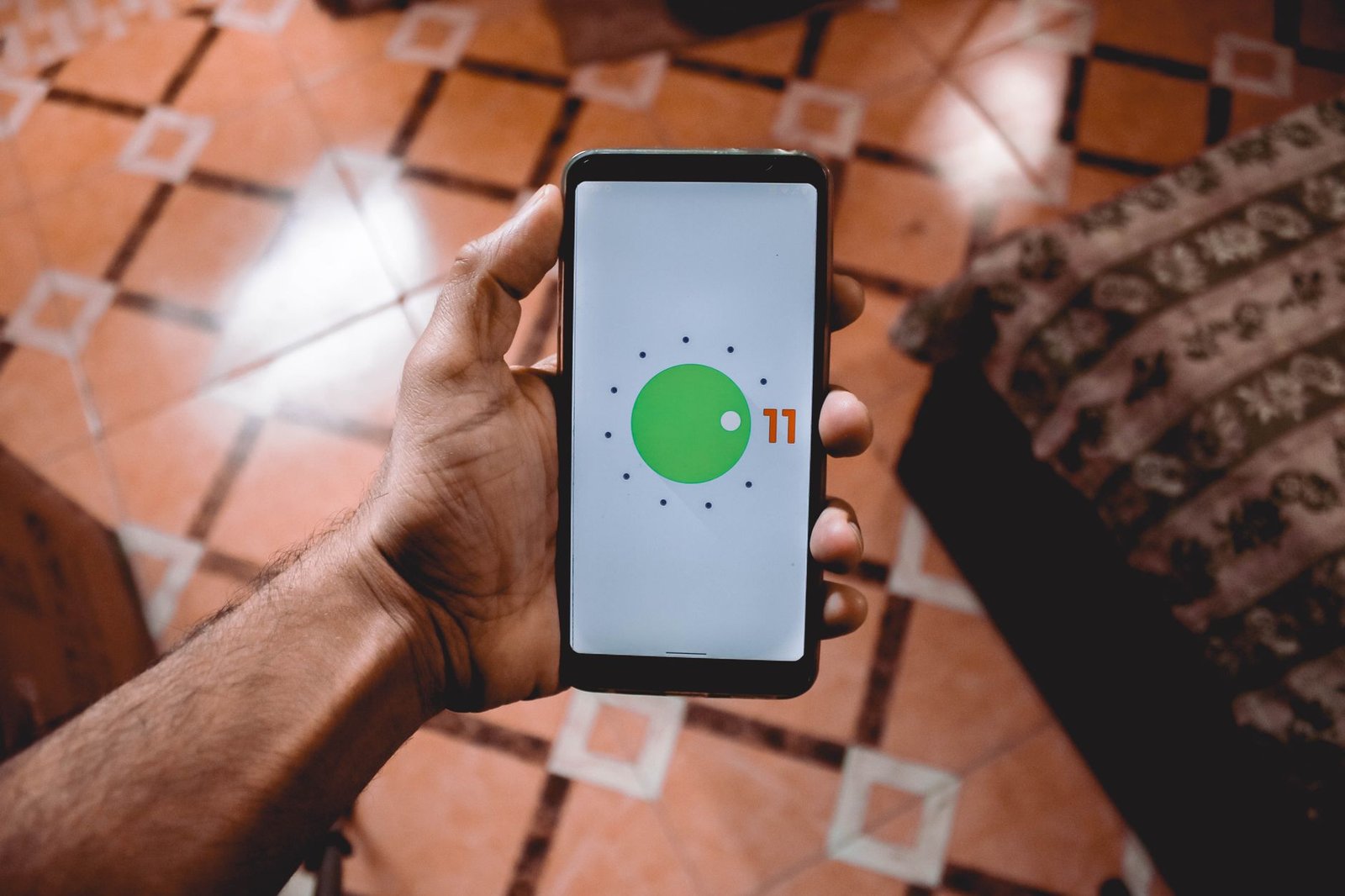 Close-up of a hand holding a smartphone displaying Android 11 interface indoors on patterned floor.