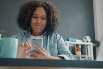 Teen girl enjoying time on smartphone in a cozy, modern room with a laptop and mug.