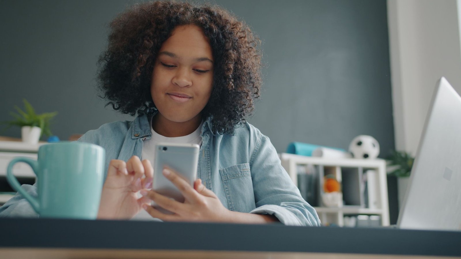 Teen girl enjoying time on smartphone in a cozy, modern room with a laptop and mug.