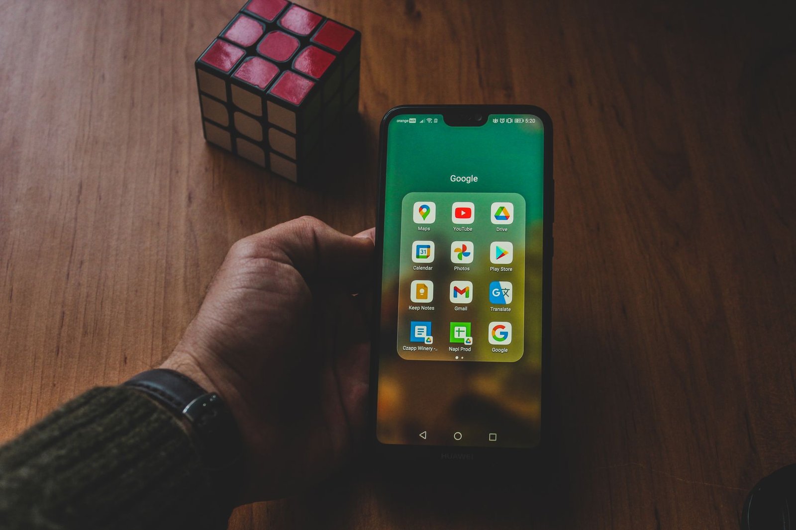 A hand holds a smartphone displaying various apps, next to a Rubik's cube on a wooden surface.