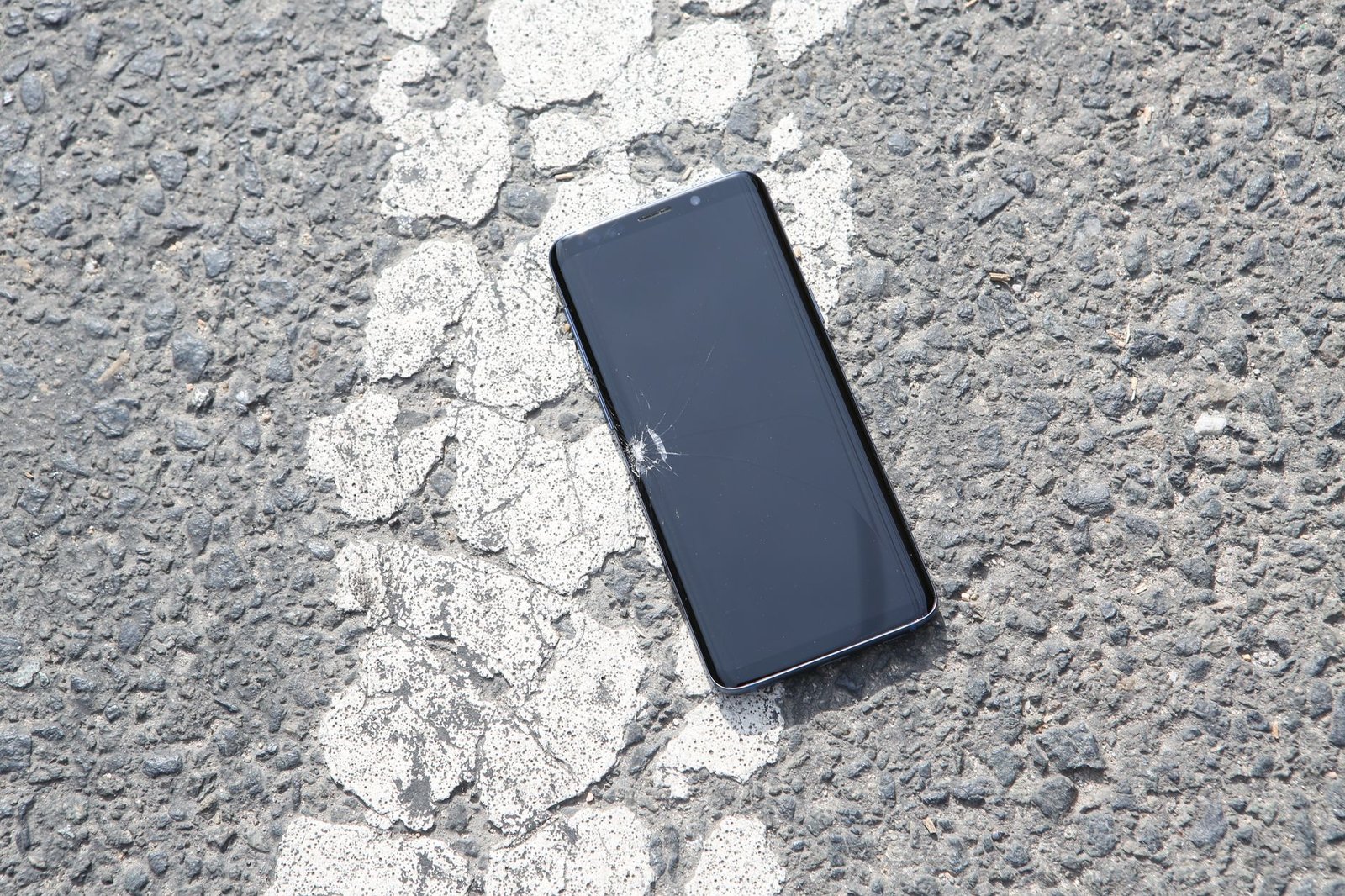 A cracked smartphone lying on a textured asphalt road surface captured from above.
