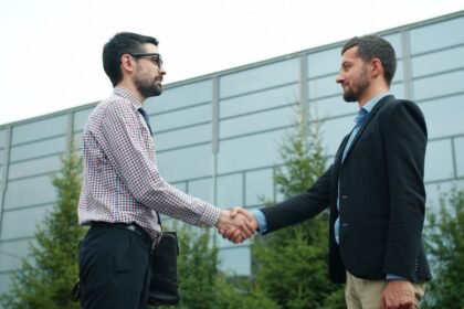 Two businessmen in casual attire shaking hands outside a modern office building.