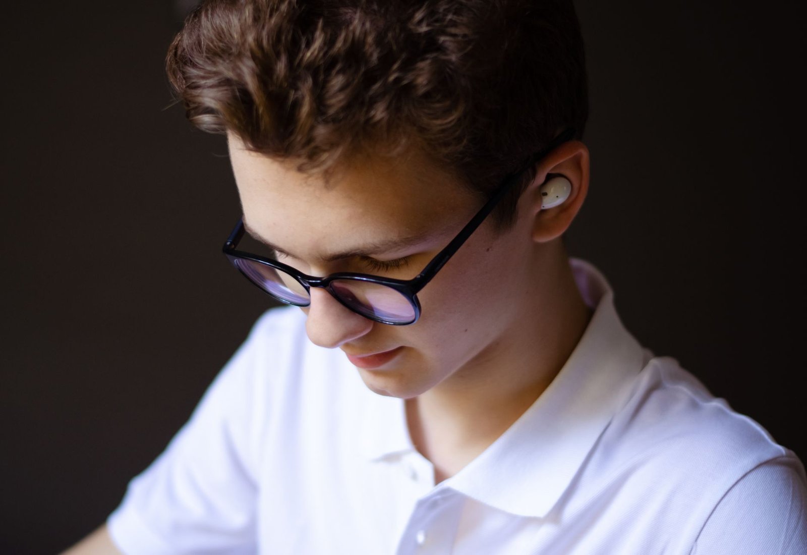Teenage boy wearing earbuds and glasses, focused on listening indoors.