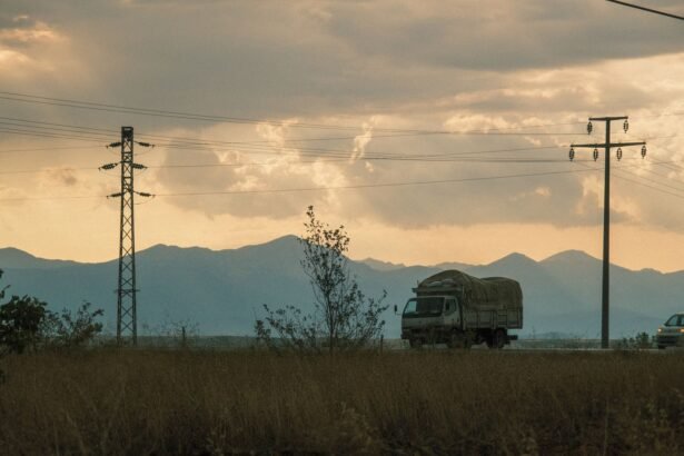 Trucks travel on a rural road at sunset, framed by power lines and distant mountains.