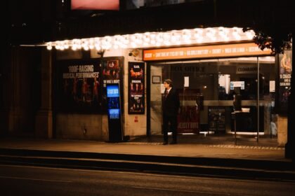 A man waits outside a brightly lit theater entrance on a city street at night, capturing urban elegance.
